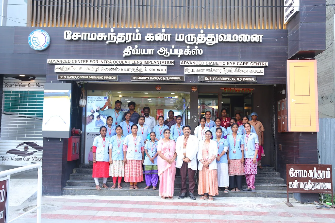 Doctors and staff at Somasundaram Eye Hospital smiling together in a bright, modern clinic environment. The group stands near medical equipment, conveying a welcoming and caring atmosphere. Wall signage reads Somasundaram Eye Hospital, emphasizing professionalism and trust.